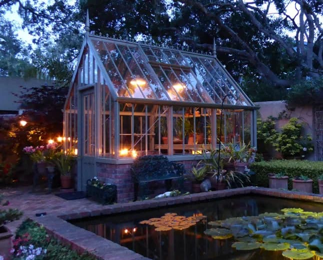 Small Alitex greenhouse in an olive finish lit from inside at dusk beside a lily pond