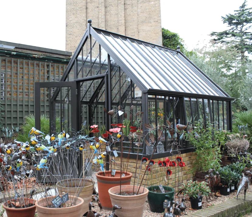 Dark grey Alitex greenhouse on a brick base surrounded by potted plants and garden ornaments