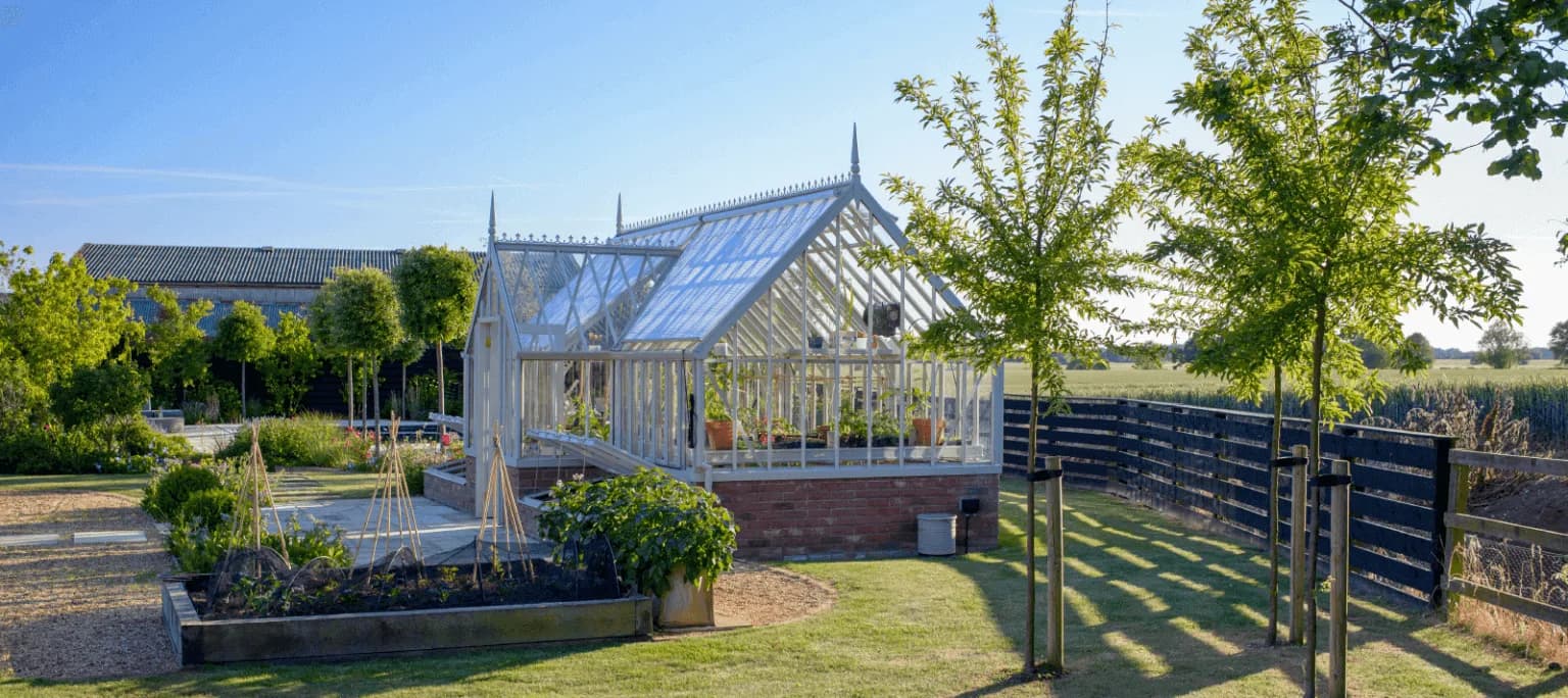 White Alitex greenhouse on a brick plinth beside raised vegetable beds in a country kitchen garden