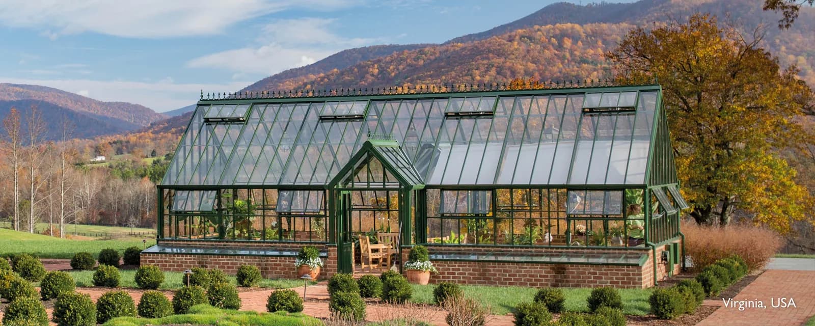 Hartley Botanic Victorian Grand Manor glasshouse in dark green on a brick dwarf wall in Virginia, with autumn mountains behind
