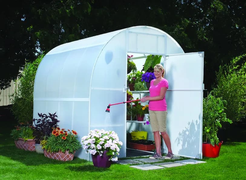 Solexx Gardener's Oasis greenhouse with the door open and a gardener watering shelves of potted flowers