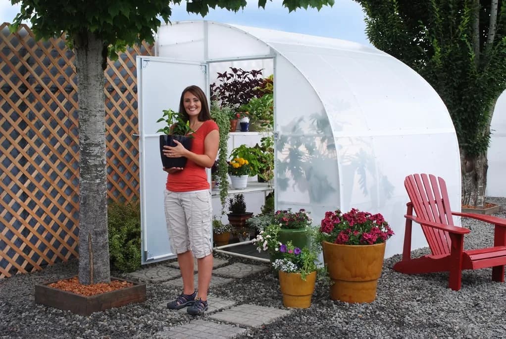 Solexx Harvester lean-to greenhouse set against a lattice wall with a gardener holding a potted plant