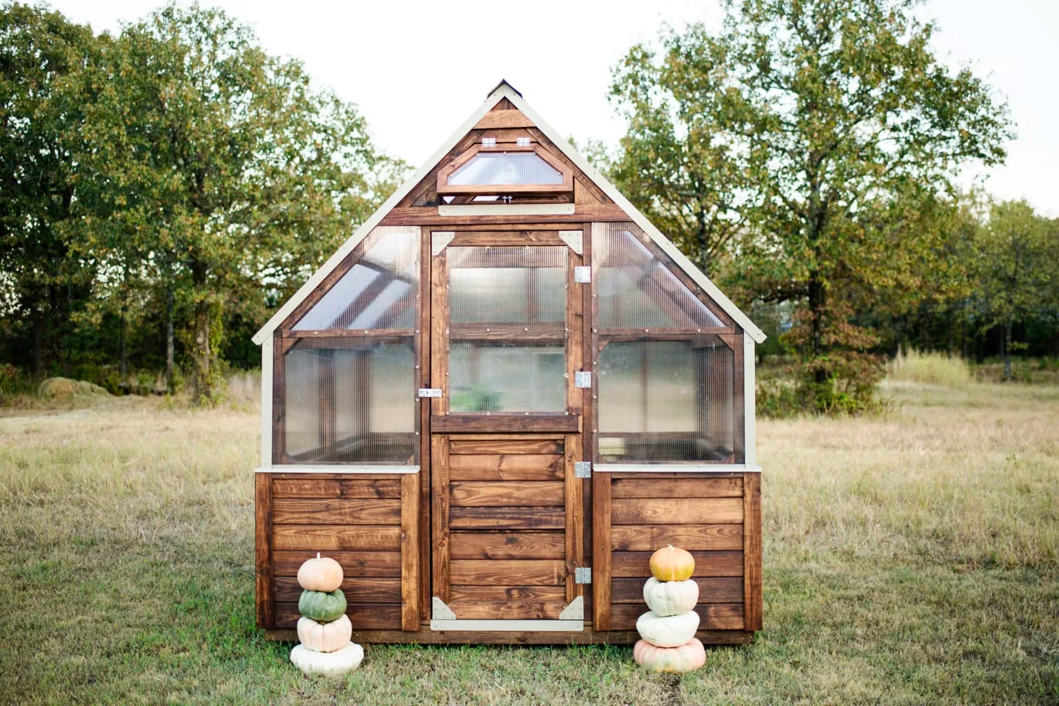 Large wooden Yoderbilt greenhouse with a steep glass roof surrounded by lush garden plantings